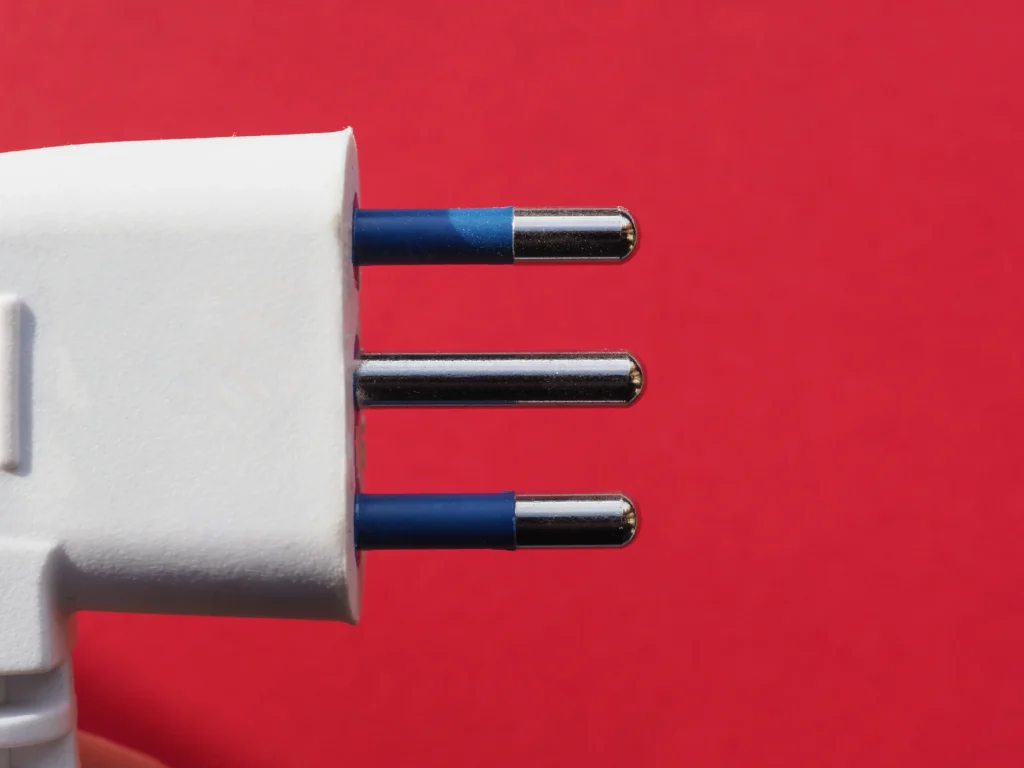Imagem de um adaptador de tomada com três pinos cilíndricos prateados, conectado a uma fonte de energia, com fundo vermelho. Conteúdo sobre plugues e tomadas.