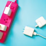 Fita extensora para tomada elétrica com três dispositivos conectados, incluindo carregadores e adaptadores, sobre fundo azul. Conteúdo sobre plugues e tomadas.