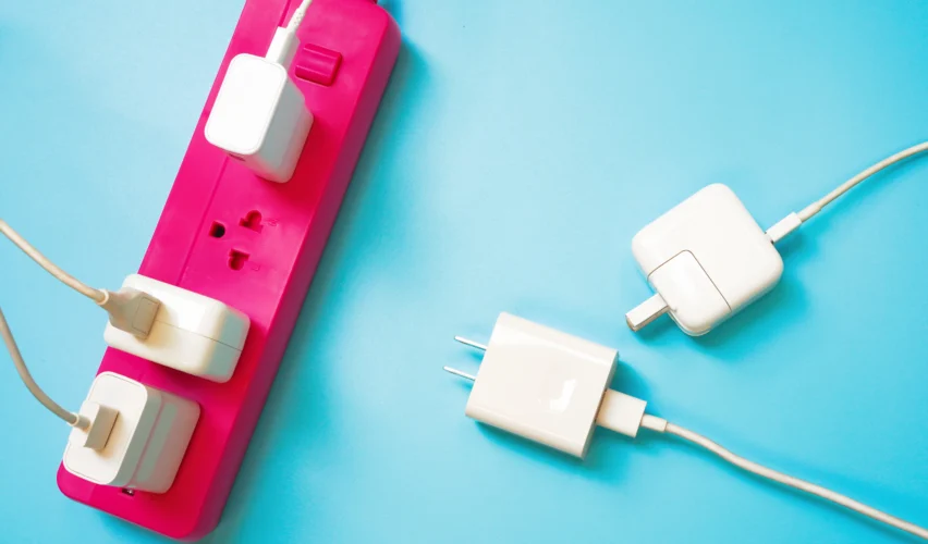 Fita extensora para tomada elétrica com três dispositivos conectados, incluindo carregadores e adaptadores, sobre fundo azul. Conteúdo sobre plugues e tomadas.