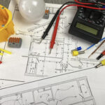 Técnico de manutenção verificando o projeto elétrico com diagramas elétricos em painel de controle de sistema industrial com componentes eletrônicos ao fundo.