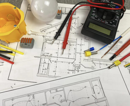 Técnico de manutenção verificando o projeto elétrico com diagramas elétricos em painel de controle de sistema industrial com componentes eletrônicos ao fundo.