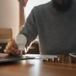 Homem com barba usando roupa cinza, sentado em uma mesa de madeira, segurando uma lâmpada e uma moeda de cobre, com pilhas de moedas ao lado. Ao lado de um laptop e smartphone, em ambiente de escritório. Conteúdo sobre como economizar energia.