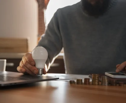 Homem com barba usando roupa cinza, sentado em uma mesa de madeira, segurando uma lâmpada e uma moeda de cobre, com pilhas de moedas ao lado. Ao lado de um laptop e smartphone, em ambiente de escritório. Conteúdo sobre como economizar energia.