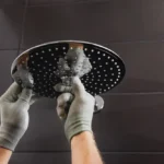 Instalação de um chuveiro Lorenzetti 220v de teto moderno em um banheiro com azulejos escuros, com mãos de trabalhador ajustando o equipamento.