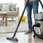 Pessoa usando aspirador de pó e água para limpeza de piso em sala de estar moderna com sofá cinza e mesa de madeira.
