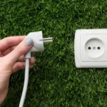 Imagem de uma mão segurando um plugue de energia próximo a uma tomada elétrica instalada em uma área com grama verde. Ideal para ilustrar dicas de energia elétrica, instalação de tomadas ou cuidados com eletricidade.
