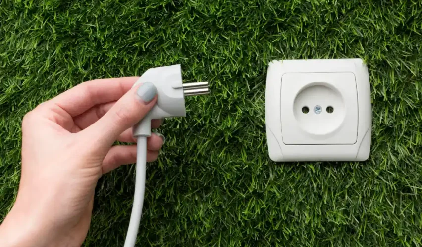 Imagem de uma mão segurando um plugue de energia próximo a uma tomada elétrica instalada em uma área com grama verde. Ideal para ilustrar dicas de energia elétrica, instalação de tomadas ou cuidados com eletricidade.