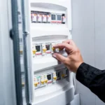 Mão ajustando e removendo disjuntor em quadro elétrico industrial com módulos e identificações, ao lado de outros componentes de proteção em painel de energia