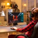 Luminárias de mesa iluminam um setup gamer aconchegante com pessoa usando computador e fones de ouvido, teclado e monitor em ambiente moderno com decoração.