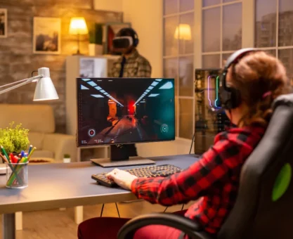 Luminárias de mesa iluminam um setup gamer aconchegante com pessoa usando computador e fones de ouvido, teclado e monitor em ambiente moderno com decoração.