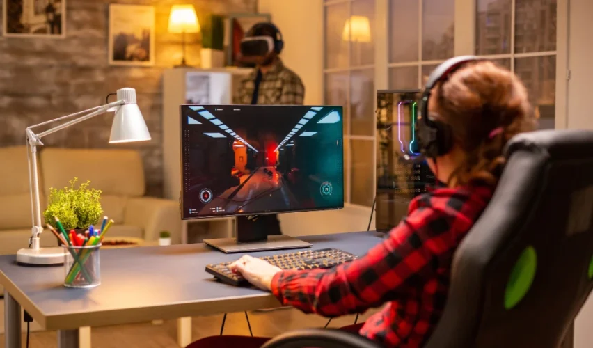 Luminárias de mesa iluminam um setup gamer aconchegante com pessoa usando computador e fones de ouvido, teclado e monitor em ambiente moderno com decoração.
