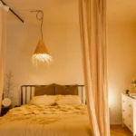 Iluminação no quarto com luminária pendente estilo cúpula de tecido sobre a cama, luz quente e cortinas claras, criando ambiente aconchegante e moderno.