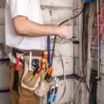 Técnico em obra elétrica na instalação de cabeamento, com ferramentas na cintura e diversos fios organizados em uma área de alvenaria, próximo a uma tabela de cabos elétricos.