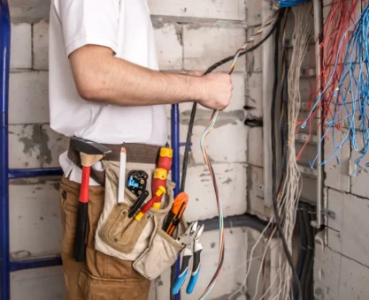 Técnico em obra elétrica na instalação de cabeamento, com ferramentas na cintura e diversos fios organizados em uma área de alvenaria, próximo a uma tabela de cabos elétricos.