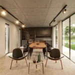 Ambiente externo moderno com mesa de jantar e poltronas, iluminação em trilho eletrificado preto no teto e refletor direcionável em fachada com grandes portas de vidro e jardim ao fundo.