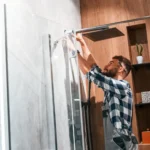Homem instalando ou ajustando o chuveiro no banheiro, com as mãos trabalhando na estrutura e preparando a troca de componentes como a resistência do chuveiro.