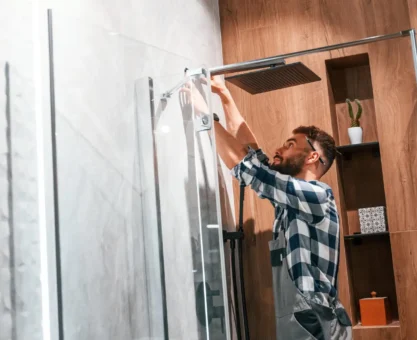 Homem instalando ou ajustando o chuveiro no banheiro, com as mãos trabalhando na estrutura e preparando a troca de componentes como a resistência do chuveiro.