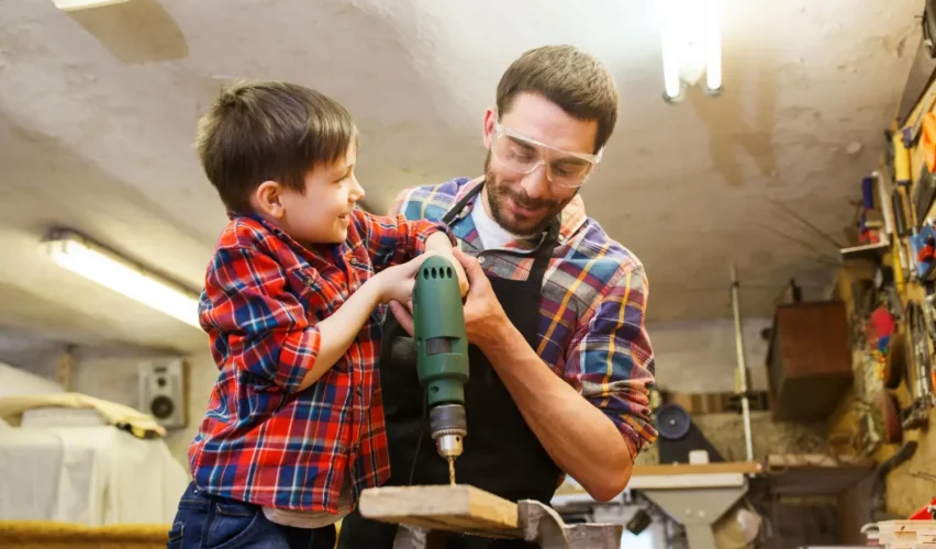 Pai e filho na oficina fazendo um trabalho de marcenaria, usando uma furadeira e peças de madeira; ideias de presentes para o dia dos pais