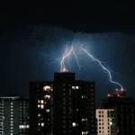 dps classe 2: Tempestade com raios intensos iluminando o céu sobre prédios e torres residenciais ao fundo, em cena noturna dramática. Refletores e janelas acendem enquanto as descargas elétricas cruzam o horizonte.
