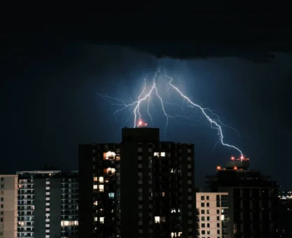 dps classe 2: Tempestade com raios intensos iluminando o céu sobre prédios e torres residenciais ao fundo, em cena noturna dramática. Refletores e janelas acendem enquanto as descargas elétricas cruzam o horizonte.