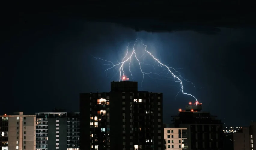 dps classe 2: Tempestade com raios intensos iluminando o céu sobre prédios e torres residenciais ao fundo, em cena noturna dramática. Refletores e janelas acendem enquanto as descargas elétricas cruzam o horizonte.