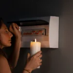 Mulher segurando uma vela acesa em casa, em frente ao disjuntor/caixa de energia, sugerindo queda de energia e falta de luz no ambiente doméstico à noite.